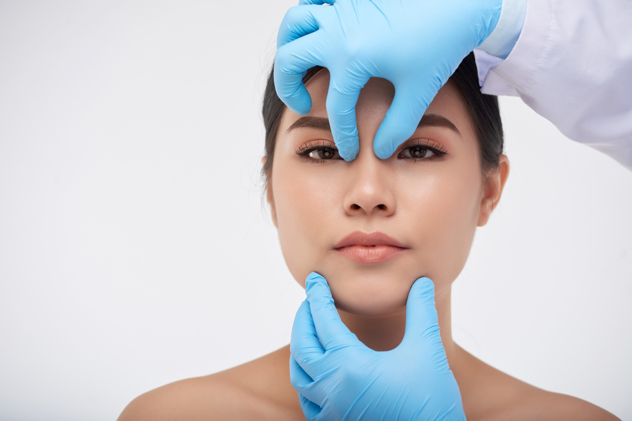 Rosto de uma mulher de cabelos pretos com as mãos de um médico avaliando nariz - motivos para escolher um especialista em rinoplastia